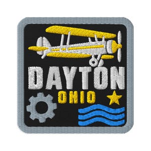 May include: A square embroidered patch with a grey border on a black background. It features a yellow and white biplane, the white text DAYTON, the yellow text OHIO, a grey gear icon, a yellow star, and three horizontal blue wavy lines.