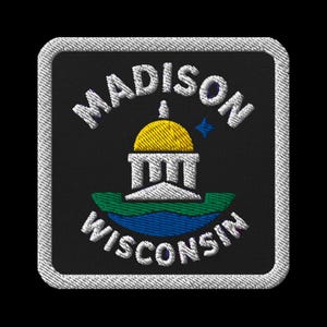 Op de afbeelding: Geborduurde patch met de woorden "MADISON" en "WISCONSIN". Het ontwerp omvat een gebouw met gele koepel, een blauwe ster en een groen en blauw landschap op een zwarte achtergrond met een witte rand.