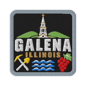 Op de afbeelding: Een vierkante geborduurde patch met een zwarte achtergrond en een grijze rand. Het toont een witte kerktoren, groene heuvels, de witte tekst GALENA, de gouden tekst ILLINOIS, gekruiste gele mijnbouwgereedschappen, blauwe golven en rode druiven.