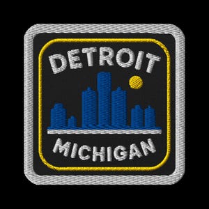 May include: Embroidered patch featuring the Detroit skyline in blue, a yellow sun, and the words "DETROIT MICHIGAN" in white. The patch has a black background with a yellow border and a white stitched edge.