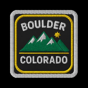 Può includere: Toppa ricamata con la scritta "BOULDER COLORADO" in bianco, sopra una catena montuosa verde con cime innevate e un sole giallo. La toppa ha uno sfondo nero con un bordo giallo e un bordo cucito bianco.