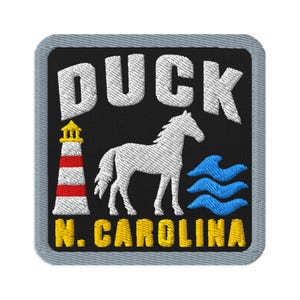 May include: A square embroidered patch with a black background and a grey border. It features the white text DUCK at the top, a white horse in the centre, a red and white lighthouse on the left, blue waves on the right, and yellow text N. CAROLINA.