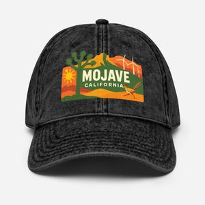 Può includere: Un cappellino da baseball nero dall'aspetto invecchiato con una grafica Mojave California. La grafica include un paesaggio desertico con un albero di Giosuè, sole, montagne, mulini a vento e il testo "Mojave California".