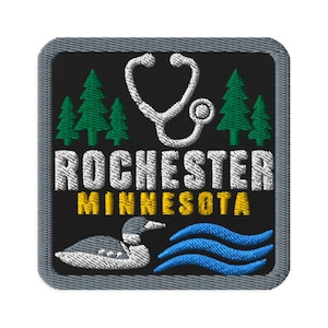 May include: A square embroidered patch with a black background and a gray border. It features a white stethoscope, green pine trees, the white text ROCHESTER, yellow text MINNESOTA, a white and gray loon, and blue wavy lines at the bottom.