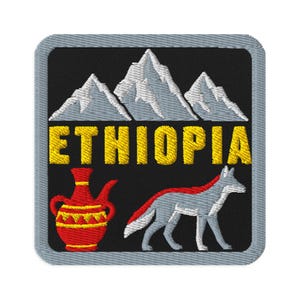 Op de afbeelding: Een vierkante geborduurde patch met een grijze rand op een zwarte achtergrond. Het toont witte bergtoppen boven de gele tekst ETHIOPIA. Onder de tekst staan een rood-gele traditionele koffiepot en een grijze en rode Ethiopische wolf.