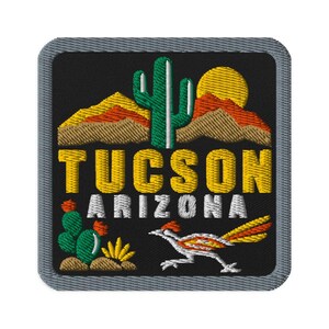 Può includere: Una toppa quadrata ricamata su sfondo nero raffigurante un cactus saguaro verde, un sole giallo e montagne marroni. Il testo TUCSON ARIZONA è ricamato in giallo e bianco. In basso sono presenti un cactus verde e un uccello corridore della strada.