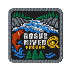 May include: A square embroidered patch with a grey border featuring a black background. The design depicts a leaping orange fish, blue river rapids, green pine trees, and a yellow fishing reel. The text reads ROGUE RIVER OREGON in white.