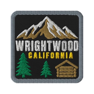 May include: A square embroidered patch with a gray border on a black background. It features a brown and white mountain range above the white text WRIGHTWOOD and yellow text CALIFORNIA, with two green pine trees and a brown log cabin below.