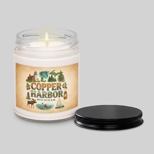 Op de afbeelding: Een brandende, witte kaars in een heldere glazen pot met een zwart deksel. Het etiket bevat de woorden "Copper Harbor Michigan" met illustraties van een vuurtoren, eland en andere buitenelementen.