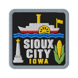 May include: A square embroidered patch with a black background and a gray border. It features a white and red steamboat with a yellow paddle wheel, a white obelisk with a yellow star, a yellow ear of corn, and the text SIOUX CITY IOWA in white and yellow.