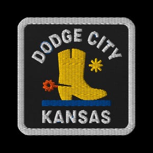 May include: Embroidered patch featuring "Dodge City Kansas" in white text. A yellow cowboy boot is centered, with a blue line beneath it. The patch has a black background and a gray border.
