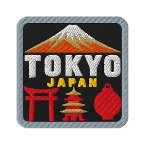 Puede incluir: Un parche bordado cuadrado con borde gris sobre fondo negro. Presenta una montaña blanca y naranja, el texto blanco TOKYO, el texto amarillo JAPAN, una puerta torii roja, una pagoda roja y dorada, y una linterna circular roja.