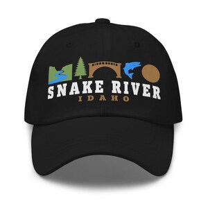 Op de afbeelding: Een zwarte baseballpet met een afbeelding van een blauwe rivier, een groene dennenboom, een bruine brug, een blauwe vis en een bruine cirkel. Onder de afbeelding staat de tekst SNAKE RIVER in het wit en IDAHO in het bruin op het voorpaneel.