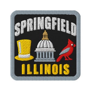 Op de afbeelding: Een vierkante geborduurde patch met een zwarte achtergrond en een grijze rand. Het bevat de witte tekst SPRINGFIELD boven een gele hoge hoed, een grijs en goudkleurig capitoolgebouw en een rode kardinaalvogel, met de gele tekst ILLINOIS eronder.