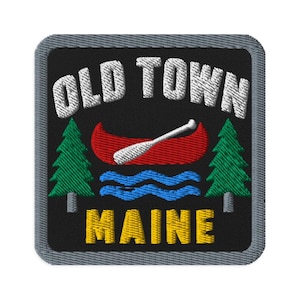 May include: A square embroidered patch with a black background and a gray border. It features the white text OLD TOWN above a red canoe with a white paddle, blue water waves, two green pine trees, and the yellow text MAINE at the bottom.