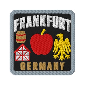 May include: A square embroidered patch with a black background and a grey border. It features the white text FRANKFURT at the top and GERMANY at the bottom. The centre displays a brown barrel, a red apple, a red and white house, and a yellow eagle.