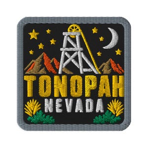 Puede incluir: Un parche bordado cuadrado con fondo negro que muestra un castillete minero blanco, montañas naranjas, estrellas amarillas y una luna creciente blanca. El texto TONOPAH es amarillo y NEVADA blanco, con plantas del desierto amarillas.