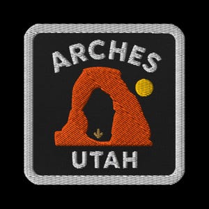 Pode incluir: Patch bordado com o texto "ARCHES UTAH" em branco, com um arco laranja e um sol amarelo sobre um fundo preto. O patch tem uma borda branca.