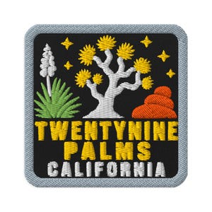 Puede incluir: Un parche bordado cuadrado con fondo negro que presenta un árbol de Josué blanco con follaje amarillo, una planta de agave verde, una flor de yuca blanca, formaciones rocosas naranjas y estrellas amarillas. El texto dice TWENTYNINE PALMS CALIFORNIA.