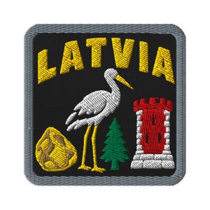 Op de afbeelding: Een vierkante geborduurde patch met een grijze rand op een zwarte achtergrond. Het toont de gele tekst LATVIA boven een witte ooievaar, een gele rots, een groene dennenboom en een rood-witte stenen toren met een gekanteelde bovenrand.