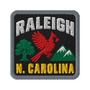 Puede incluir: Un parche bordado cuadrado con fondo negro y borde gris. Presenta el texto blanco RALEIGH en la parte superior, un pájaro cardenal rojo en el centro, un árbol verde a la izquierda, montañas verdes a la derecha y el texto amarillo N. CAROLINA.