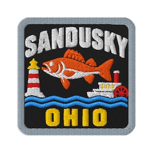 Puede incluir: Un parche bordado cuadrado con fondo negro que presenta el texto blanco SANDUSKY en la parte superior y OHIO en amarillo en la parte inferior. El diseño incluye un pez naranja, un faro rojo y blanco, y un barco de vapor blanco sobre olas azules.