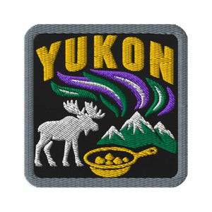 Könnte beinhalten: Ein quadratischer gestickter Aufnäher mit schwarzem Hintergrund und grauem Rand. Er zeigt den gelben Schriftzug YUKON über einem weißen Elch, grünen Bergen mit weißen Gipfeln und einer gelben Pfanne mit Essen sowie violetten und grünen Nordlichtern.