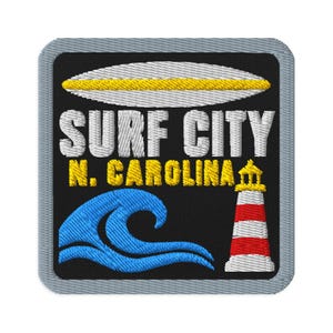 Puede incluir: Un parche bordado cuadrado con fondo negro y borde gris. Presenta una tabla de surf blanca con una franja amarilla, el texto blanco SURF CITY, el texto amarillo N. CAROLINA, una ola azul y un faro con rayas rojas y blancas.