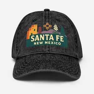 Könnte beinhalten: Schwarze Baseballkappe im Vintage-Stil mit einer farbenfrohen Grafik von Santa Fe, New Mexico. Das Design umfasst eine südwestliche Landschaft mit Gebäuden, einem Sonnensymbol und dem Text "SANTA FE NEW MEXICO" in einem grünen Banner.