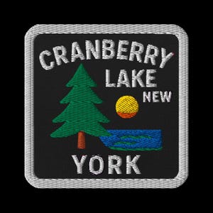 May include: Embroidered patch featuring the words "CRANBERRY LAKE NEW YORK" in white, with a green tree, yellow sun, and blue lake. The patch has a black background and a white border.