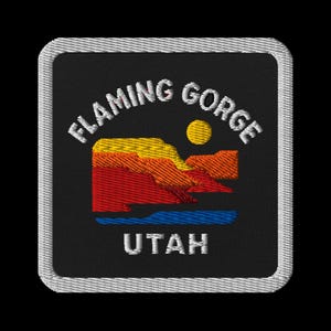 May include: Embroidered patch featuring the words "FLAMING GORGE" above a landscape scene with a yellow sun, and the word "UTAH" below. The landscape depicts red, orange, and yellow rock formations and blue water. The patch has a black background and a white border.