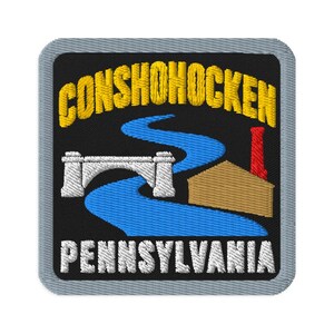 Puede incluir: Un parche bordado cuadrado con fondo negro y borde gris. Presenta el texto amarillo CONSHOHOCKEN sobre un río azul, un puente de piedra blanco, un edificio color canela con una chimenea roja y el texto blanco PENNSYLVANIA debajo.