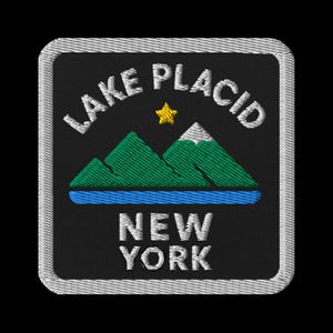 May include: Embroidered patch featuring the words "Lake Placid New York". The design includes green mountains, a blue lake, and a yellow star. The patch has a black background and a white border.