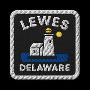 以下が含まれることがあります： 黒い背景に白い文字で「LEWES DELAWARE」と書かれた刺繍パッチ。デザインは、黄色い上部と黄色の太陽が付いた白い灯台が特徴です。パッチには白い縁取りがあります。