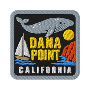 Puede incluir: Un parche bordado cuadrado con fondo negro que muestra una ballena gris, un velero blanco, un sol amarillo y acantilados naranjas sobre agua azul. El texto DANA POINT está en amarillo y la palabra CALIFORNIA está en blanco abajo.