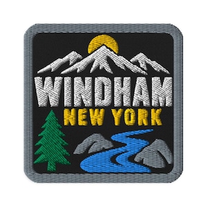 May include: A square embroidered patch with a black background and a gray border. It features white mountain peaks under a yellow sun, the white text WINDHAM, yellow text NEW YORK, a green pine tree, a blue river, and gray rocks.