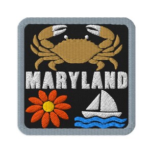 Könnte beinhalten: Ein quadratischer gestickter Aufnäher mit grauem Rand auf schwarzem Hintergrund. Er zeigt eine beige Krabbe über dem weißen Schriftzug MARYLAND. Unter dem Text befinden sich eine orangefarbene Blume mit gelber Mitte und ein weißes Segelboot auf blauem Wellenwasser.