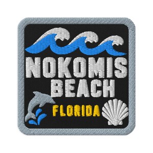 Puede incluir: Un parche bordado cuadrado con fondo negro que presenta olas azules en la parte superior, texto blanco NOKOMIS BEACH, texto amarillo FLORIDA, una silueta de delfín gris a la izquierda y una concha marina blanca en la parte inferior derecha.