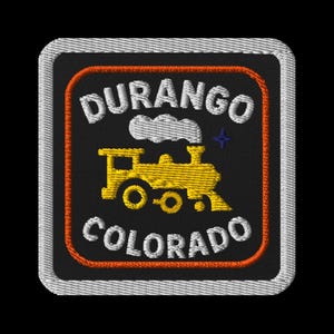 May include: Embroidered patch featuring a yellow train with the text "DURANGO COLORADO" in white lettering. The patch has a black background with an orange border and a white stitched edge. A blue star is visible.