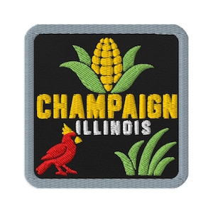 Op de afbeelding: Een vierkante geborduurde patch met een zwarte achtergrond en een grijze rand. Het toont een gele maïskolf met groene bladeren, de gele tekst "CHAMPAIGN", de witte tekst "ILLINOIS", een rode kardinaalvogel en groene grassprieten.