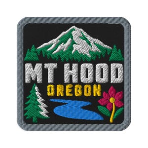 Puede incluir: Un parche bordado cuadrado con fondo negro que muestra una montaña blanca nevada, pinos verdes y un río azul. El texto MT HOOD está en blanco y OREGON en amarillo. A la derecha hay una flor rosa con el centro de color amarillo.