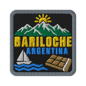 Può includere: Una toppa ricamata quadrata con sfondo nero che raffigura un sole giallo che sorge sopra montagne verdi e bianche. Il testo recita BARILOCHE in giallo e ARGENTINA in blu. Sotto si trovano una barca a vela bianca su onde blu e una tavoletta di cioccolato marrone.