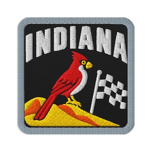 May include: A square embroidered patch with a grey border on a black background. It features the white text INDIANA arched at the top, a red cardinal bird standing on a yellow and orange mound, and a white and black chequered racing flag.