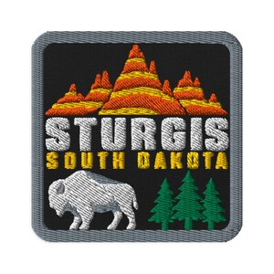 May include: A square embroidered patch with a black background featuring orange and yellow mountain peaks at the top. The text STURGIS in white and SOUTH DAKOTA in yellow appear below. A white buffalo and three green pine trees are at the bottom.