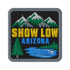 May include: A square embroidered patch with a gray border on a black background. It features a mountain range with white snow-capped peaks and green trees. The text SHOW LOW is in yellow, ARIZONA is in blue, with a green pine tree and a blue lake.
