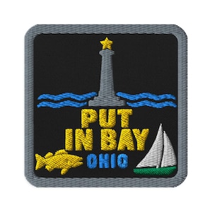 Puede incluir: Un parche bordado cuadrado con fondo negro y borde gris. Presenta un faro gris con una estrella amarilla, olas azules, un pez amarillo, un velero blanco con casco verde, y el texto amarillo PUT IN BAY y el texto azul OHIO.