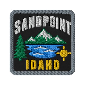 May include: A square embroidered patch with a black background and a gray border. It features the white text SANDPOINT arched above a green mountain range with white peaks, a blue lake, a green pine tree, a yellow sun, and the yellow text IDAHO.