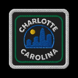 Puede incluir: Parche bordado con las palabras "Charlotte" y "Carolina" en blanco, encima y debajo de un horizonte azul y un sol amarillo. El parche tiene un fondo negro con bordes verdes y blancos.