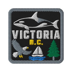 May include: A square embroidered patch with a black background and a gray border. It features a white orca, the text "VICTORIA" in white, "B.C." in yellow, a gray eagle, a white sailboat on blue waves, and a green pine tree.