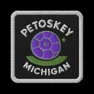May include: Embroidered patch with the text "PETOSKEY MICHIGAN" in white, above and below a purple football design. A green curved line is beneath the ball. The patch has a black background and a white stitched border.